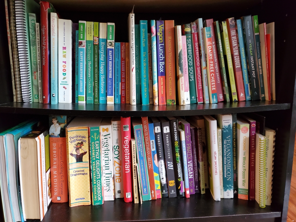 A bookshelf with two full rows of cookbooks. The bowed top shelf has books on raw foods, a set of Happy Herbivore books, vegan dessert books, vegetarian  cookbooks for kids, and other vegan books. The bottom shelf has large vegetarian cookbooks and miscellaneous vegan cookbooks.  
