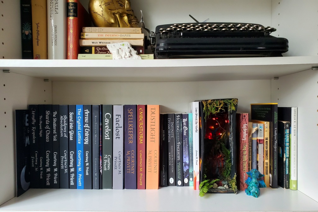 Two shelves of a white bookcase. The top shelf has a vintage typewriter, and some classic literature both vertically and stacked horizontally. On the horizontal pile is a golden skull.
The lower shelf has the complete works of Courtney M. Privett (22 books, including anthologies) filling 2/3 of the shelf, and a mossy book nook and several books by Ray Bradbury and Ursula LeGuin filling the rest. There is a tiny blue dragon sitting in front of those books.