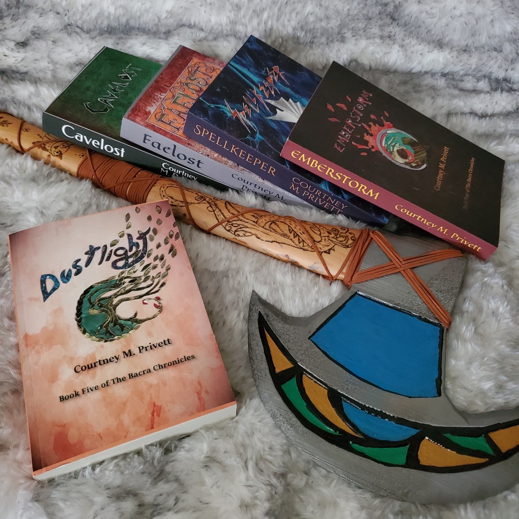 Five paperback books and a colorful battle axe sit on a furry surface. The books are Cavelost, Faelost, Spellkeeper, Emberstorm, and Dustlight. They are the complete series of The Bacra Chronicles, by Courtney M. Privett. The axe has a moon-shaped blade that is abstract patterned with blue, green, yellow, and black inlays. The handle has images carved into it, the most visible being a stylized dragon.